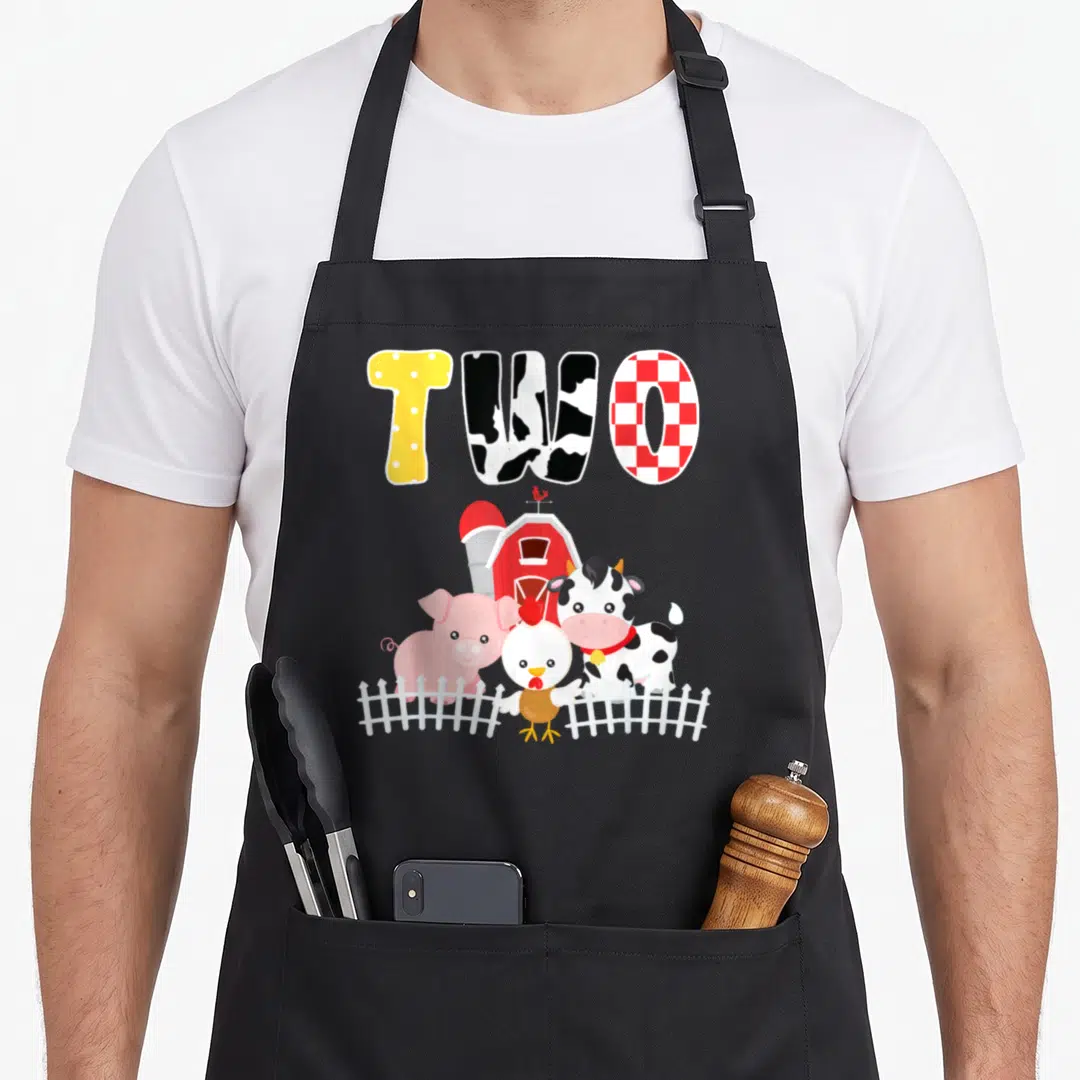 Barnyard animal farm aprons - Cartoon cow pig chicken behind white fence under patterned word TWO — shown on a black cotton canvas apron against a white background