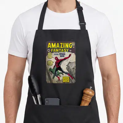 Cover book apron - Retro comic cover with yellow Amazing Fantasy title and red blue Spider-Man swinging above grey — shown on a black cotton canvas apron against a white background