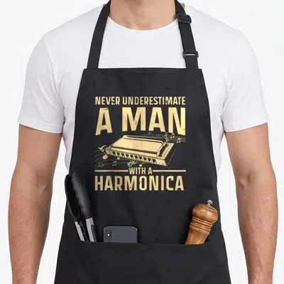 Instrument harmonica apron - Tan distressed text NEVER UNDERESTIMATE A MAN WITH A HARMONICA surrounding a harmonica — shown on a black cotton canvas apron against a white background