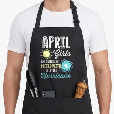 April sunshine birthday apron - Stacked white yellow blue text reading APRIL Girls Are Sunshine Mixed With A Little — shown on a black cotton canvas apron against a white background