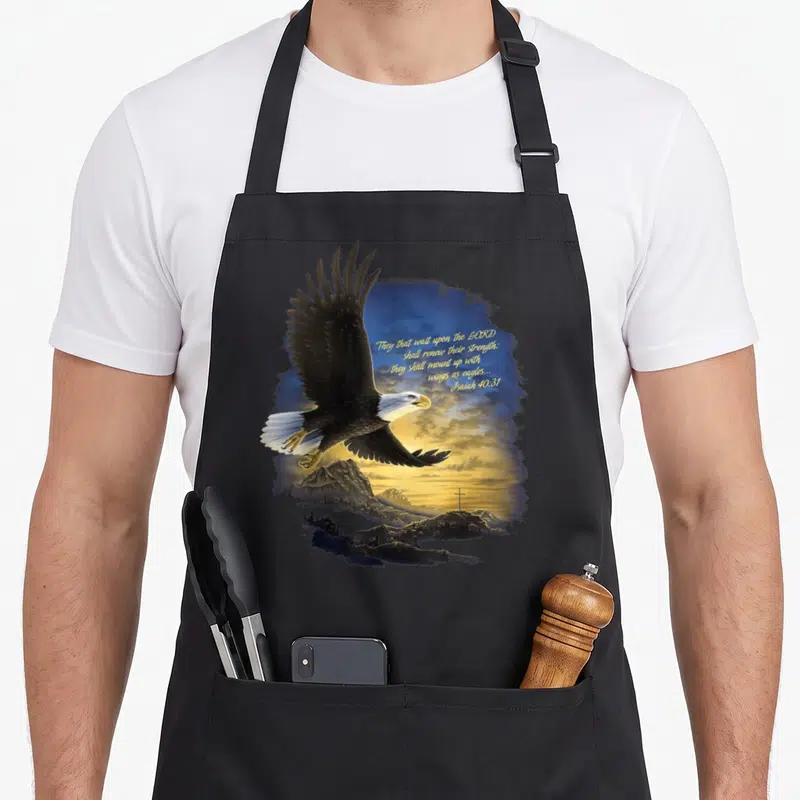 Wing bible verse apron - Realistic eagle flying over mountains with a cross and yellow Isaiah 40:31 text on blue sky — shown on a black cotton canvas apron against a white background