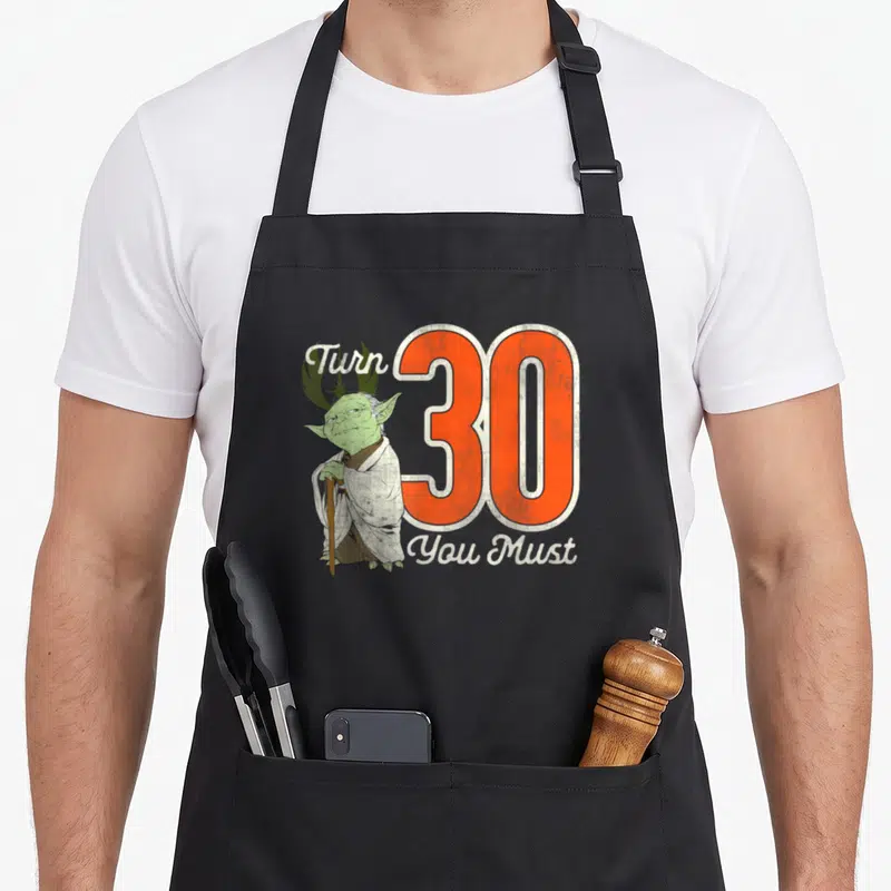 Yoda 30th birthday apron - Cartoon Yoda standing beside a large orange number 30 with white script text — shown on a black cotton canvas apron against a white background