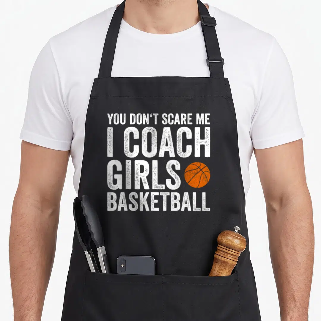 Coach panic attack apron - Stacked white text YOU DON'T SCARE ME I COACH GIRLS BASKETBALL with orange basketball graphic — shown on a black cotton canvas apron against a white background