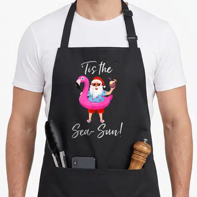 Flamingo santa claus apron - Cartoon Santa in pink flamingo float holding coconut drink with text Tis the Sea-Sun — shown on a black cotton canvas apron against a white background
