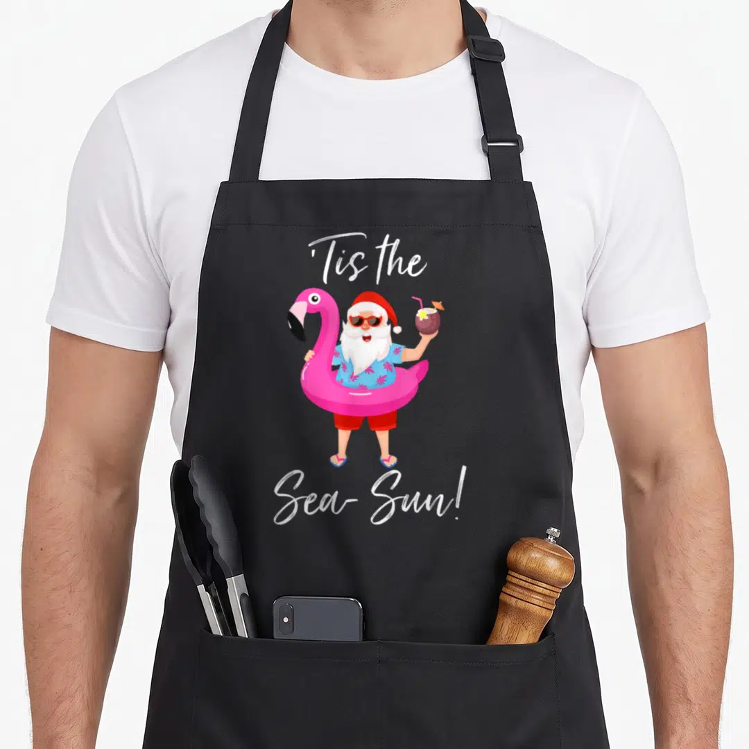 Flamingo santa claus apron - Cartoon Santa in pink flamingo float holding coconut drink with text Tis the Sea-Sun — shown on a black cotton canvas apron against a white background