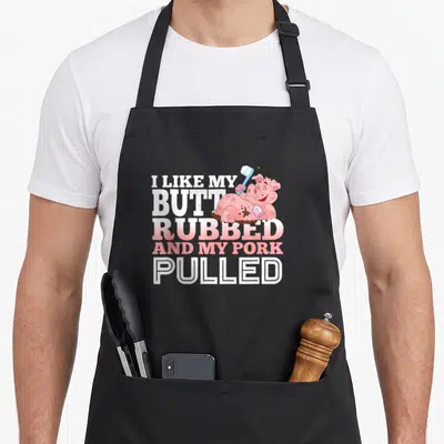 Pulled funny pig barbecue backyard apron - Cartoon pig in mud holding toothbrush, text I LIKE MY BUTT RUBBED AND MY — shown on a black cotton canvas apron against a white background