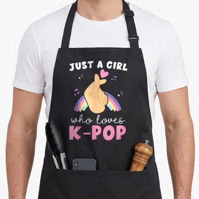 Funny pop girl apron - Cartoon hand making finger heart with rainbow and notes under Just A Girl Who Loves K-Pop text — shown on a black cotton canvas apron against a white background