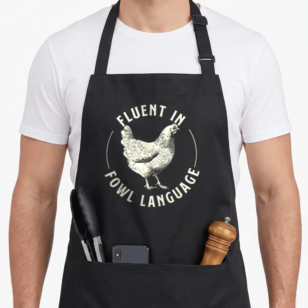 Fluent fowl language apron - White line-art chicken inside circle with arched text reading fluent in fowl language — shown on a black cotton canvas apron against a white background