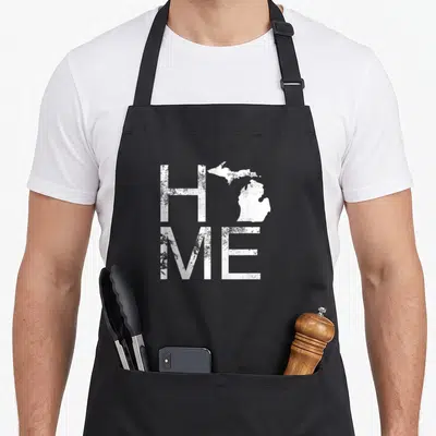 Michigan jersey pride apron - Distressed white HOME text with Michigan map replacing the O on a black background — shown on a black cotton canvas apron against a white background