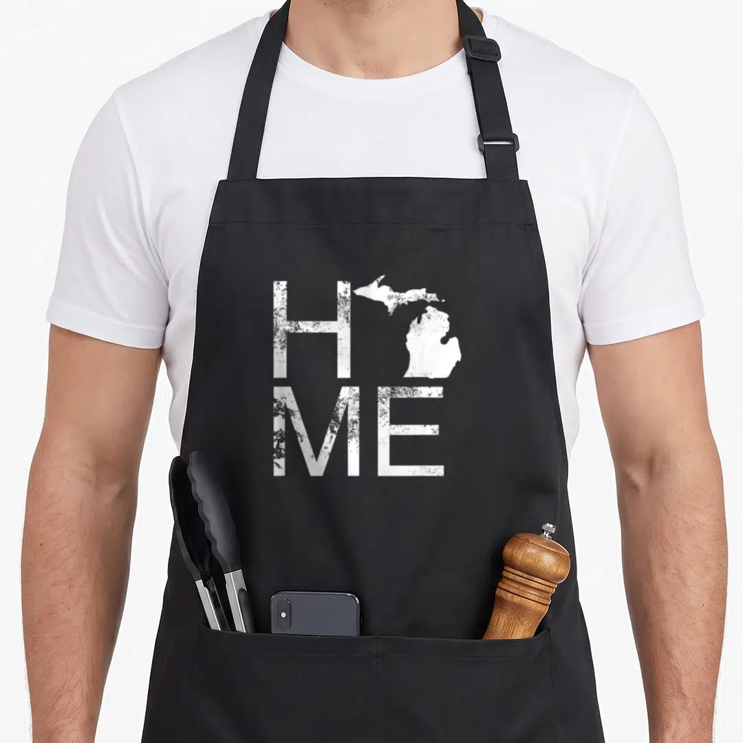 Michigan jersey pride apron - Distressed white HOME text with Michigan map replacing the O on a black background — shown on a black cotton canvas apron against a white background