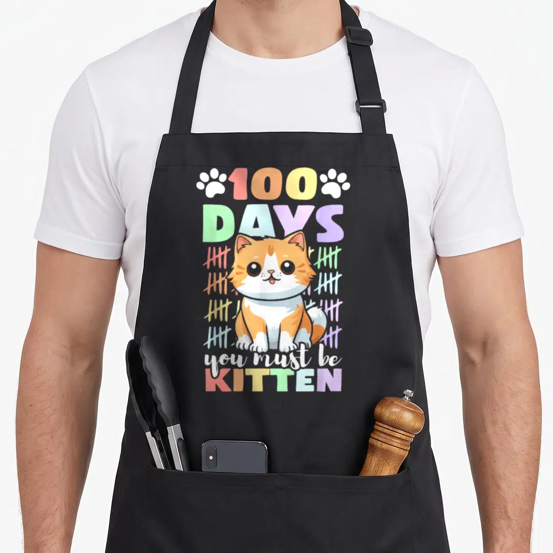 100th cat apron - Cartoon orange and white kitten surrounded by colorful tally marks and 100 DAYS text — shown on a black cotton canvas apron against a white background