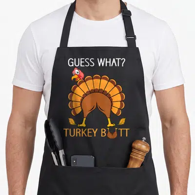 Fuuny thanksgiving apron - Cartoon turkey from behind with GUESS WHAT? TURKEY BUTT text and two fall leaves — shown on a black cotton canvas apron against a white background