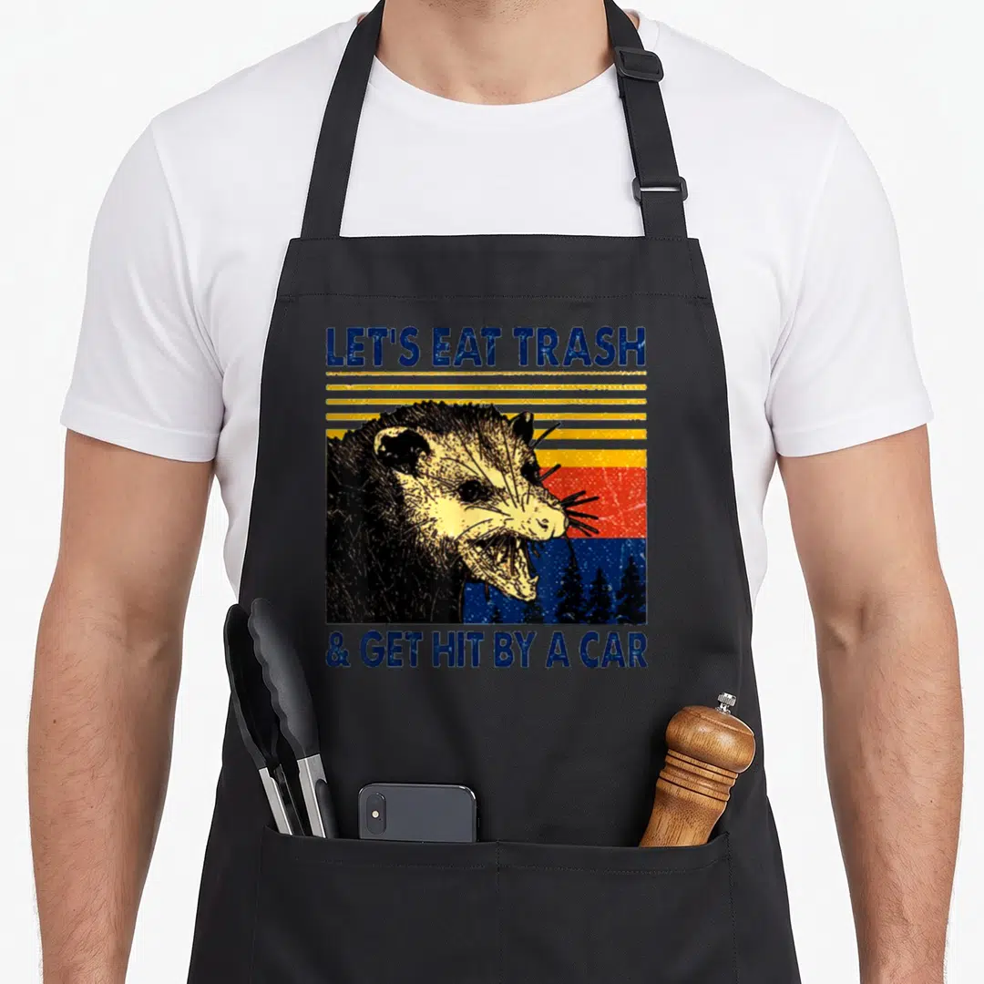 Trash let's eat apron - Retro opossum head against blue yellow orange and red sunset stripes with bold blue text — shown on a black cotton canvas apron against a white background