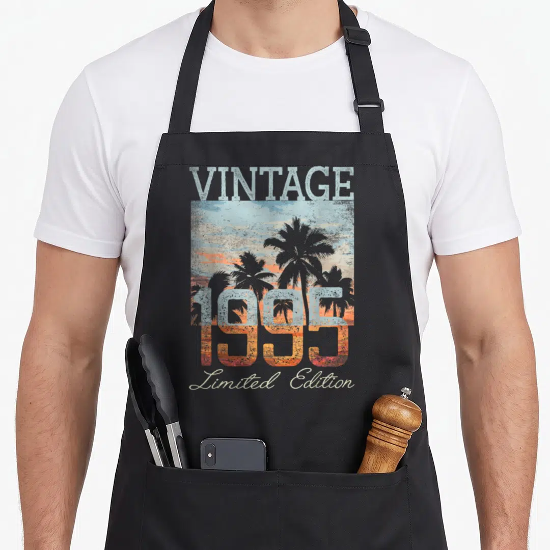 Edition 31st birthday apron - Vintage 1995 Limited Edition text over retro palm tree sunset scene in grey blue orange — shown on a black cotton canvas apron against a white background