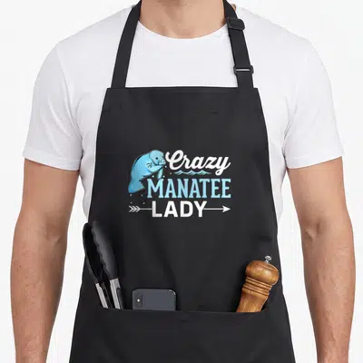 Manatee sea cow apron - Cartoon blue manatee beside stacked white and blue text with waves and arrow underline — shown on a black cotton canvas apron against a white background