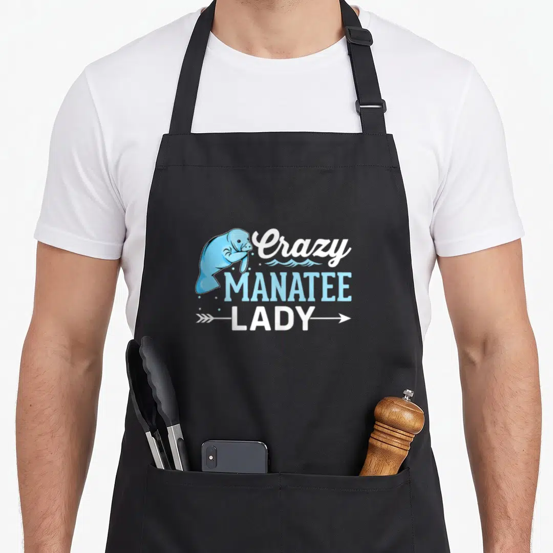 Manatee sea cow apron - Cartoon blue manatee beside stacked white and blue text with waves and arrow underline — shown on a black cotton canvas apron against a white background