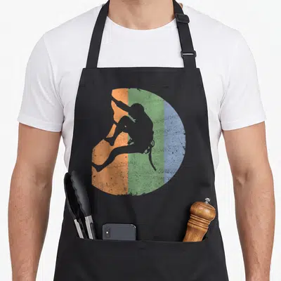 Rock outdoor apron - Grey climber silhouette over orange, green, and blue vertical bands in a circle — shown on a black cotton canvas apron against a white background