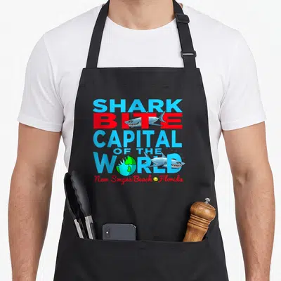 Shark daytona apron - Stacked text reading Shark Bite Capital of the World with cartoon sharks and globe in blue and red — shown on a black cotton canvas apron against a white background