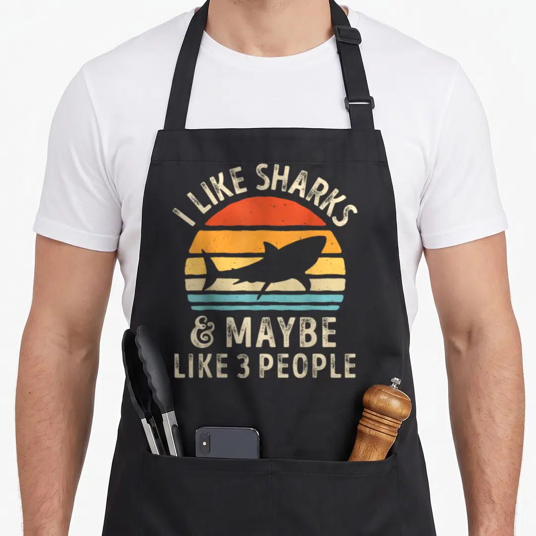 Maybe shark lover apron - Retro striped sun with centered shark silhouette and sarcastic quote text — shown on a black cotton canvas apron against a white background
