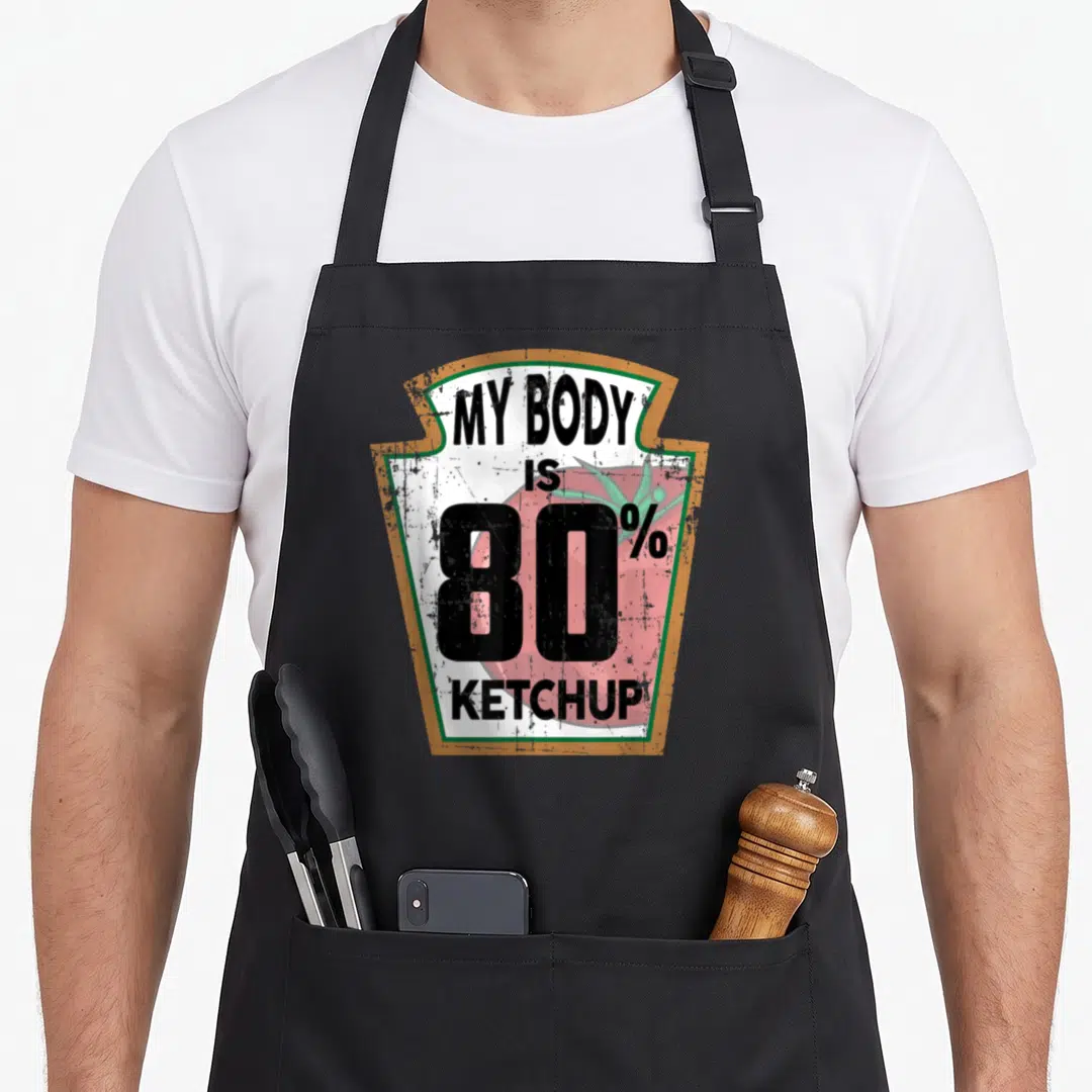 Themed joke apron - Shield containing red tomato and text MY BODY IS 80% KETCHUP in distressed black font — shown on a black cotton canvas apron against a white background