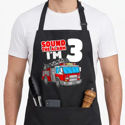 Firefighter truck apron - SOUND THE ALARM I'M 3 text with red white grey fire truck cartoon — shown on a black cotton canvas apron against a white background