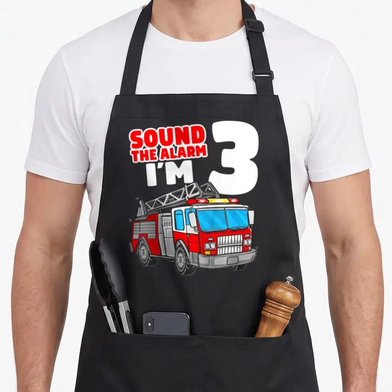 Firefighter truck apron - SOUND THE ALARM I'M 3 text with red white grey fire truck cartoon — shown on a black cotton canvas apron against a white background