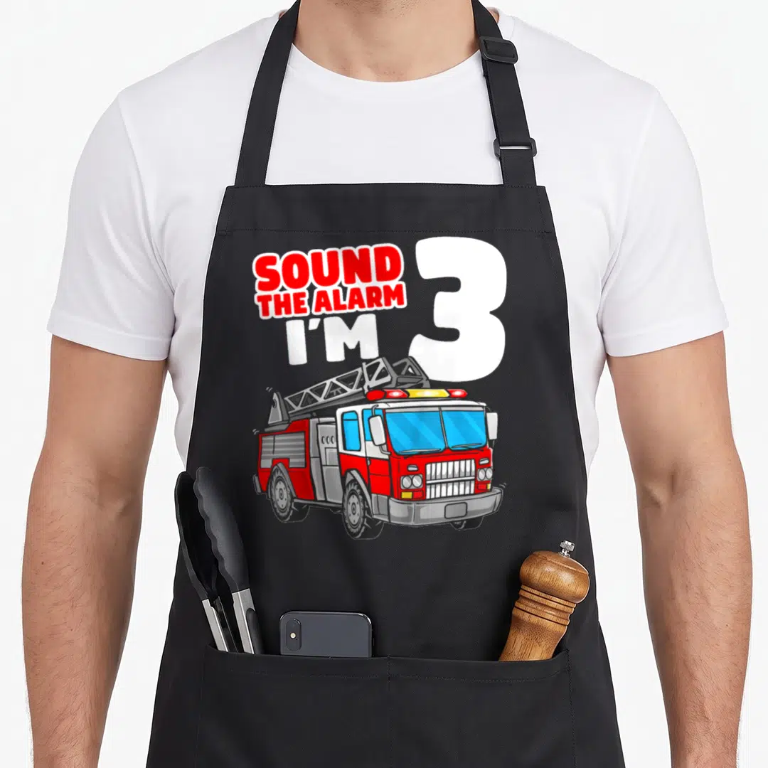 Firefighter truck apron - SOUND THE ALARM I'M 3 text with red white grey fire truck cartoon — shown on a black cotton canvas apron against a white background