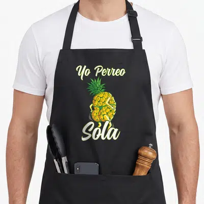 Sola pineapple apron - Dancing yellow pineapple with Yo Perreo Sola text in cartoon style — shown on a black cotton canvas apron against a white background