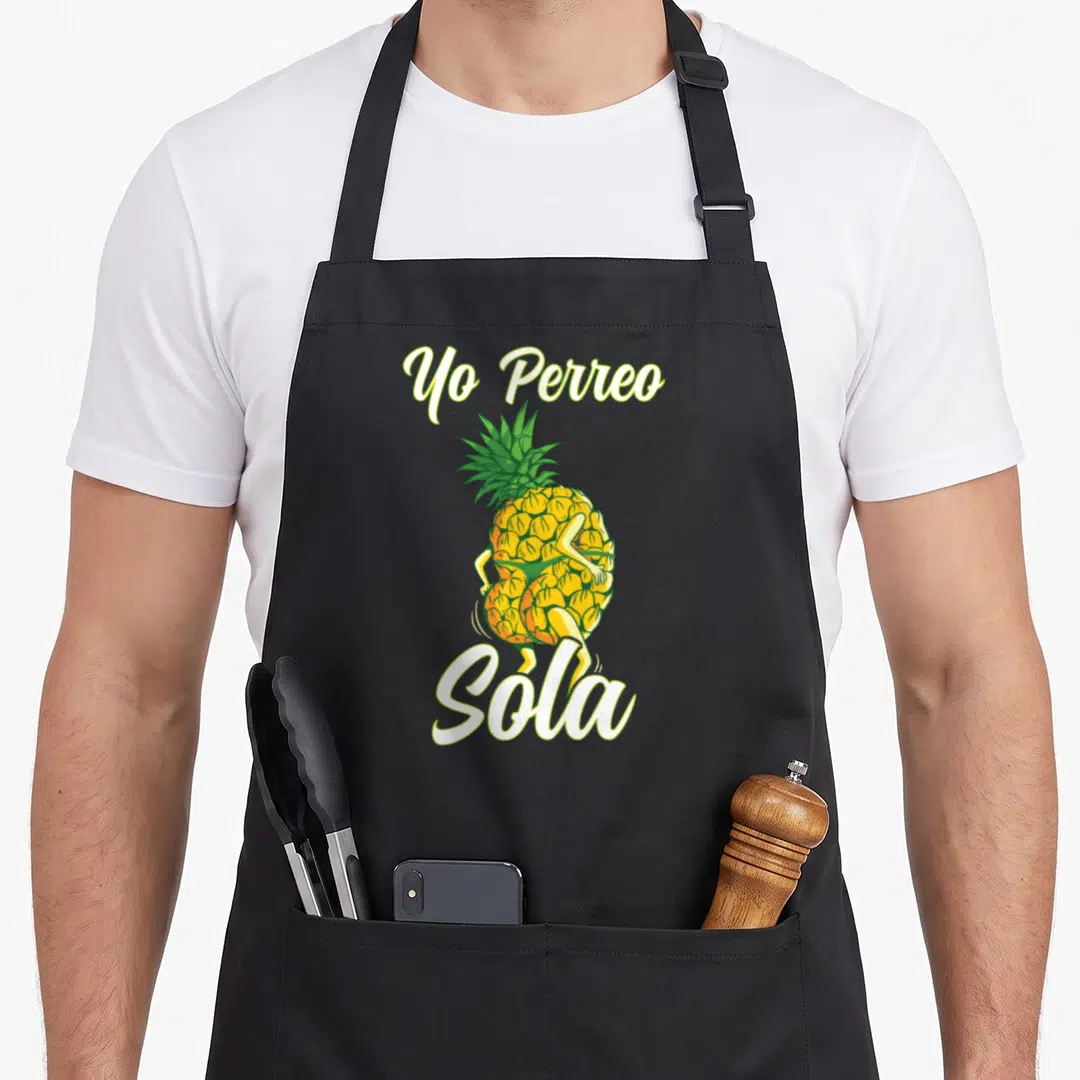 Sola pineapple apron - Dancing yellow pineapple with Yo Perreo Sola text in cartoon style — shown on a black cotton canvas apron against a white background