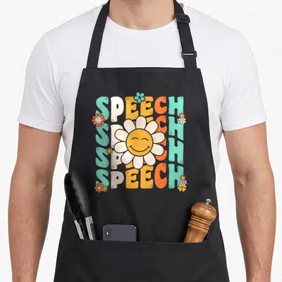 Language pathologist my therapy apron - Stacked SPEECH text with smiling white daisy and small orange teal flowers — shown on a black cotton canvas apron against a white background