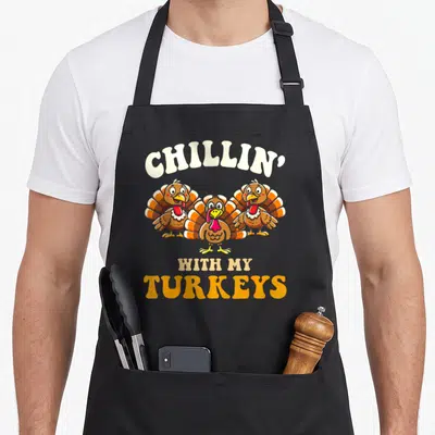 Chillin thanksgiving apron - Three cartoon turkeys with arched cream text and stacked orange lettering reading CHILLIN — shown on a black cotton canvas apron against a white background