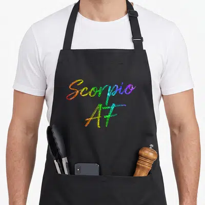 October november birthday apron - Scorpio AF stacked rainbow glitter text in red orange yellow green blue purple — shown on a black cotton canvas apron against a white background