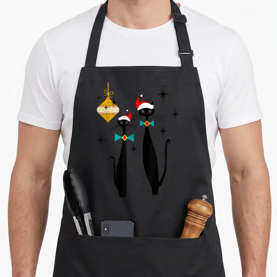 Christmas modern apron - Two black cats in Santa hats and teal bow ties beside a yellow geometric ornament and black — shown on a black cotton canvas apron against a white background