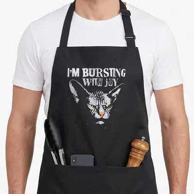 Bursting cat apron - I'M BURSTING WITH JOY text above a grey Sphynx cat head with yellow eyes — shown on a black cotton canvas apron against a white background