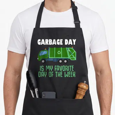 Trash truck apron - Green cartoon garbage truck with text GARBAGE DAY IS MY FAVORITE DAY OF THE WEEK — shown on a black cotton canvas apron against a white background