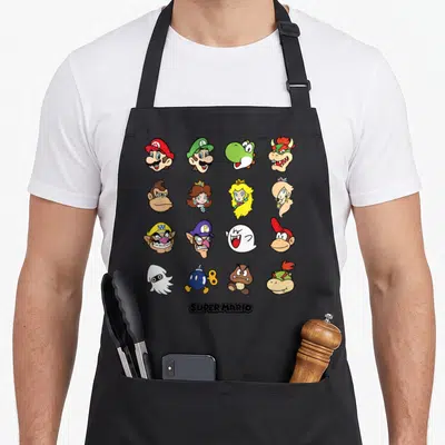Face holiday apron - Sixteen cartoon character heads in a 4x4 grid with SUPER MARIO text below — shown on a black cotton canvas apron against a white background