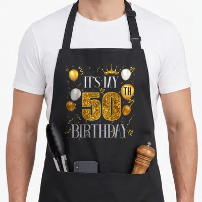50th bday apron - Glittery gold 50 with silver IT'S MY BIRTHDAY text, gold crown, white and gold balloons, confetti — shown on a black cotton canvas apron against a white background