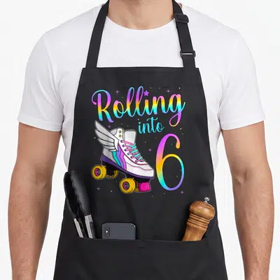 Vintage rolling turning apron - Winged roller skate with rainbow Rolling into 6 text and scattered stars — shown on a black cotton canvas apron against a white background