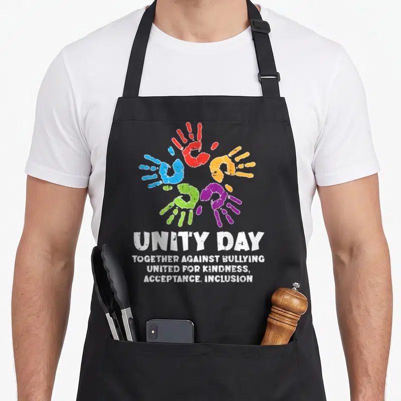 Together unity day apron - Five colorful handprints in a circle above white text reading Unity Day Together Against — shown on a black cotton canvas apron against a white background
