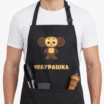 Cheburashka russian apron - Brown cartoon monkey character above tan Cyrillic text CHEBURASHKA — shown on a black cotton canvas apron against a white background
