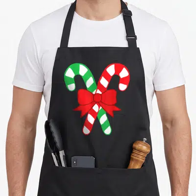 Bow candy cane apron - Crossed red and green candy canes tied with a red bow in cartoon style — shown on a black cotton canvas apron against a white background