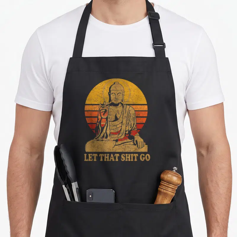 Buddha distressed apron - Retro Buddha inside yellow orange red striped circle with text LET THAT SHIT GO — shown on a black cotton canvas apron against a white background