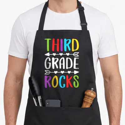 Rock art teacher aprons - Stacked THIRD GRADE ROCKS text with heart arrows in green red purple blue yellow white — shown on a black cotton canvas apron against a white background