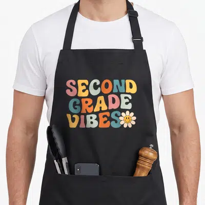 Second 1st apron 2nd apron - Stacked pink yellow blue orange white text reading SECOND GRADE VIBES with smiling daisy — shown on a black cotton canvas apron against a white background