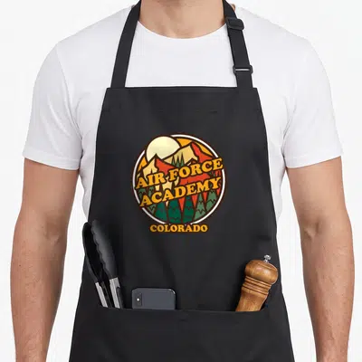 Academy colorado apron - Retro circular emblem with mountains trees and AIR FORCE ACADEMY COLORADO text — shown on a black cotton canvas apron against a white background