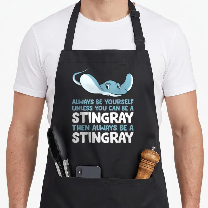 Alway stingray apron - Cartoon light blue stingray above stacked text reading Always Be Yourself Unless You Can Be — shown on a black cotton canvas apron against a white background