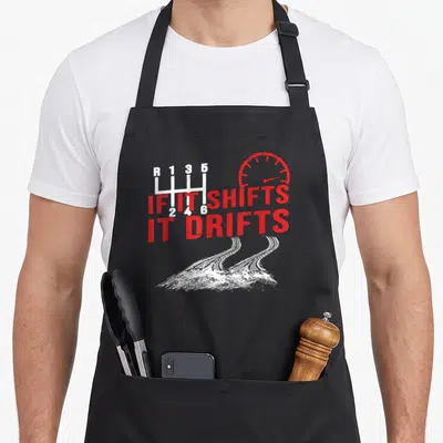 Modern car apron - Distressed red and white gear shifter speedometer and tire tracks with IF IT SHIFTS IT DRIFTS text — shown on a black cotton canvas apron against a white background