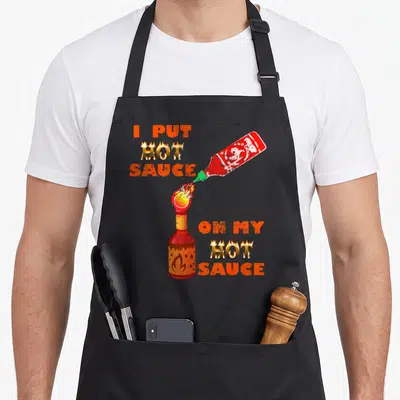 Mixed food aprons - Flaming text I PUT HOT SAUCE ON MY HOT SAUCE with two pouring bottles — shown on a black cotton canvas apron against a white background
