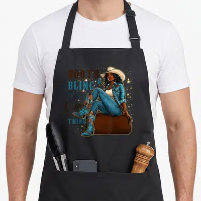 Bling cowgirl western apron - Cartoon cowgirl sitting on suitcase with boots and bling text and gold stars — shown on a black cotton canvas apron against a white background