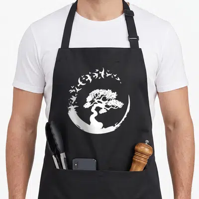 Japanese enso tree apron - White bonsai tree inside circular arc of flying birds, minimalist design on black apron — shown on a black cotton canvas apron against a white background
